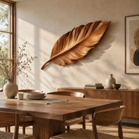 Leaf shaped wooden wall sculpture with raised carved veins and a curling stem beside a dining table in natural light