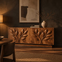 Sideboard with two carved leafy branches across four doors on a plinth in a dark dining room at golden hour