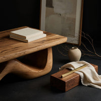 Corner detail of light toned coffee table with a wave shaped base beside wood, brass and linen samples