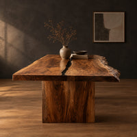 Live edge dining table with a dark center seam and a slab base in a dark studio against a charcoal plaster wall