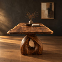 Live edge dining table on a single sculpted pedestal with a teardrop opening in a dark studio against a charcoal plaster wall