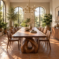 Live edge dining table on a single sculpted pedestal with a teardrop opening in a bright dining room with arched windows and plants