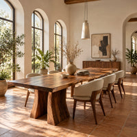Dining table with a thick live edge slab top on two flared pedestal legs in a bright dining room with arched windows and plants