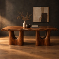 Long dining table with a live edge top and two arched pedestal legs in a dark studio against a charcoal plaster wall