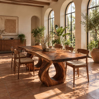 Rectangular dining table with a live edge slab top and two loop pedestals in a bright dining room with arched windows and plants