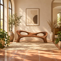 Long wood bench with a pinched middle and two oval openings in a bright entryway with arched windows and plants
