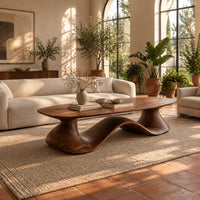 Long oval coffee table on a flowing wave shaped wooden pedestal base in a bright living room with arched windows and plants