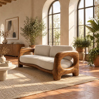 Sofa with a sculpted wood frame, open loop arms and a long seat cushion in a bright living room with arched windows and plants
