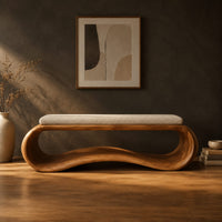 Wooden bench made from a looped ribbon frame with a slim cream cushion in a dark studio against a charcoal plaster wall