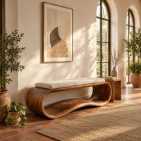 Wooden bench made from a looped ribbon frame with a slim cream cushion in a bright entryway with arched windows and plants