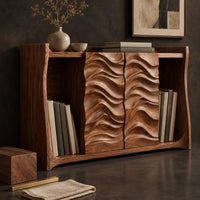 Corner detail of low bookshelf with two carved wave doors and open bays at both ends beside wood, brass and linen samples