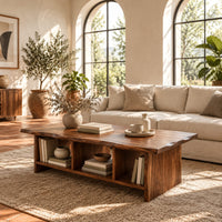 Low coffee table with a wavy natural edge top and three open cubbies in a bright living room with arched windows and plants