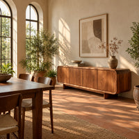 Low wood sideboard with four flat front panels and short legs in a bright dining room with arched windows and plants