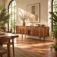 Long sideboard with four doors carved with nested arches on tapered legs in a bright dining room with arched windows and plants