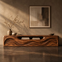 Corner detail of low wood tv stand with a rippling carved front and an open top slot beside wood, brass and linen samples
