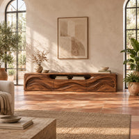 Low wood tv stand with a rippling carved front and an open top slot in a bright living room with arched windows and plants