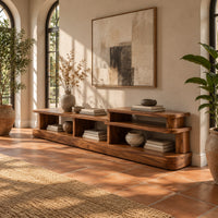 Low stepped open shelf console table with a raised section at one end in a bright entryway with arched windows and plants