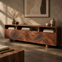 Corner detail of low wood tv stand with a carved wave front and open top compartments beside wood, brass and linen samples