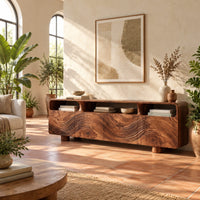 Low wood tv stand with a carved wave front and open top compartments in a bright living room with arched windows and plants