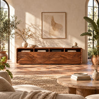 Low tv stand with four open cubbies above wave carved front panels in a bright living room with arched windows and plants
