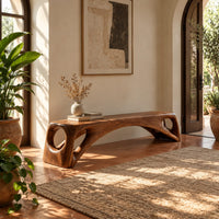 Low solid wood bench with an arched underside and open cutouts at each end in a bright entryway with arched windows and plants