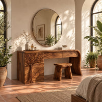 Makeup vanity with a carved tree pedestal and a sweeping slab leg in a bright bedroom with arched windows and plants