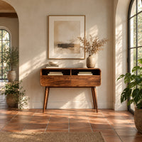 Mid century wooden console table with open compartments above two front panels in a bright entryway with arched windows and plants