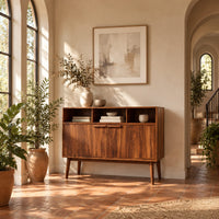 Mid century console table with three open cubbies and two doors on tapered legs in a bright entryway with arched windows and plants