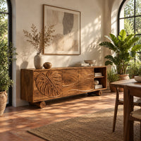 Sideboard with a carved monstera leaf door and an open end shelf in a bright dining room with arched windows and plants