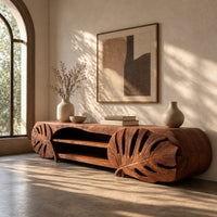 Corner detail of long tv stand with monstera leaf carved ends and open shelves in the center beside wood, brass and linen samples