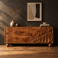 Sideboard with a carved monstera leaf on the right doors and ball feet in a dark studio against a charcoal plaster wall