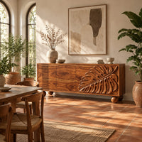 Sideboard with a carved monstera leaf on the right doors and ball feet in a bright dining room with arched windows and plants