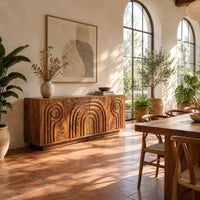 Sideboard with nested arch carving and spiral motifs on four doors in a bright dining room with arched windows and plants