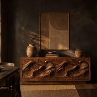 Low sideboard with four carved wave relief panels on a plinth base in a dark dining room at golden hour