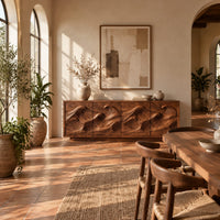 Low sideboard with four carved wave relief panels on a plinth base in a bright dining room with arched windows and plants