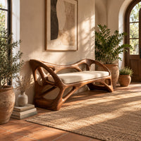 Wooden bench with a pale cushioned seat and an organic open frame in a bright entryway with arched windows and plants