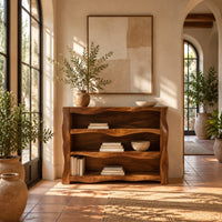 Open shelf console table with wavy sculpted sides in a bright entryway with arched windows and plants