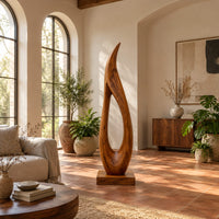 Tall teardrop sculpture with an open center and a pointed tip in a bright living room with arched windows and plants