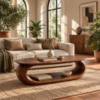 Oval coffee table on a thick pill shaped loop base with an open middle in a bright living room with arched windows and plants