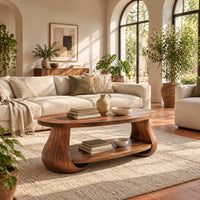 Oval coffee table with swelling tulip shaped legs and a lower shelf in a bright living room with arched windows and plants