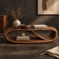 Corner detail of oval coffee table with a twisting ribbon loop body and an open lower level beside wood, brass and linen samples