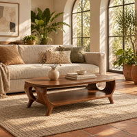 Oval coffee table with a lower shelf and flared legs with circular cutouts in a bright living room with arched windows and plants
