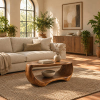 Oval coffee table with a sculpted body and open lower shelf in a bright living room with arched windows and plants