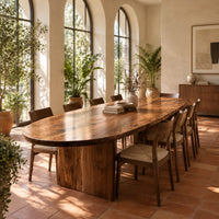 Oval dining table with a thick edge and two broad slab legs in a bright dining room with arched windows and plants