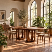Oval dining table on two pedestal bases with tall arched openings in a bright dining room with arched windows and plants
