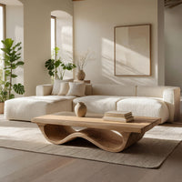 Pale hardwood coffee table with an undulating base in a sunlit room with large windows and plants