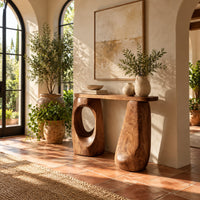 Console table with two chunky pebble shaped legs and a slim oval ended top in a bright entryway with arched windows and plants