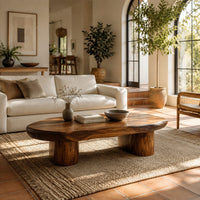 Pebble shaped coffee table with a smooth organic top and two thick column legs in a bright living room with arched windows and plants