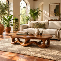 Low oval coffee table with a wavy base cut through by irregular openings in a bright living room with arched windows and plants