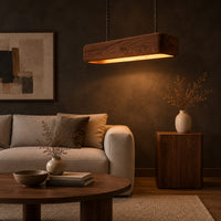 Long wood beam pendant light with soft corners and a warm glow below in a dark living room at golden hour
